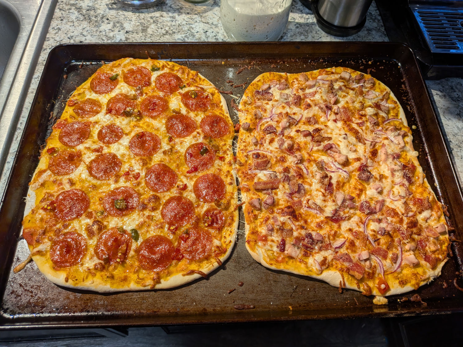 Two baked tavern-style sourdough pizzas side by side on a sheet pan, one topped with pepperoni and diced cowboy candy jalapenos, the other with smoked chicken thighs, bacon, and thinly sliced red onion