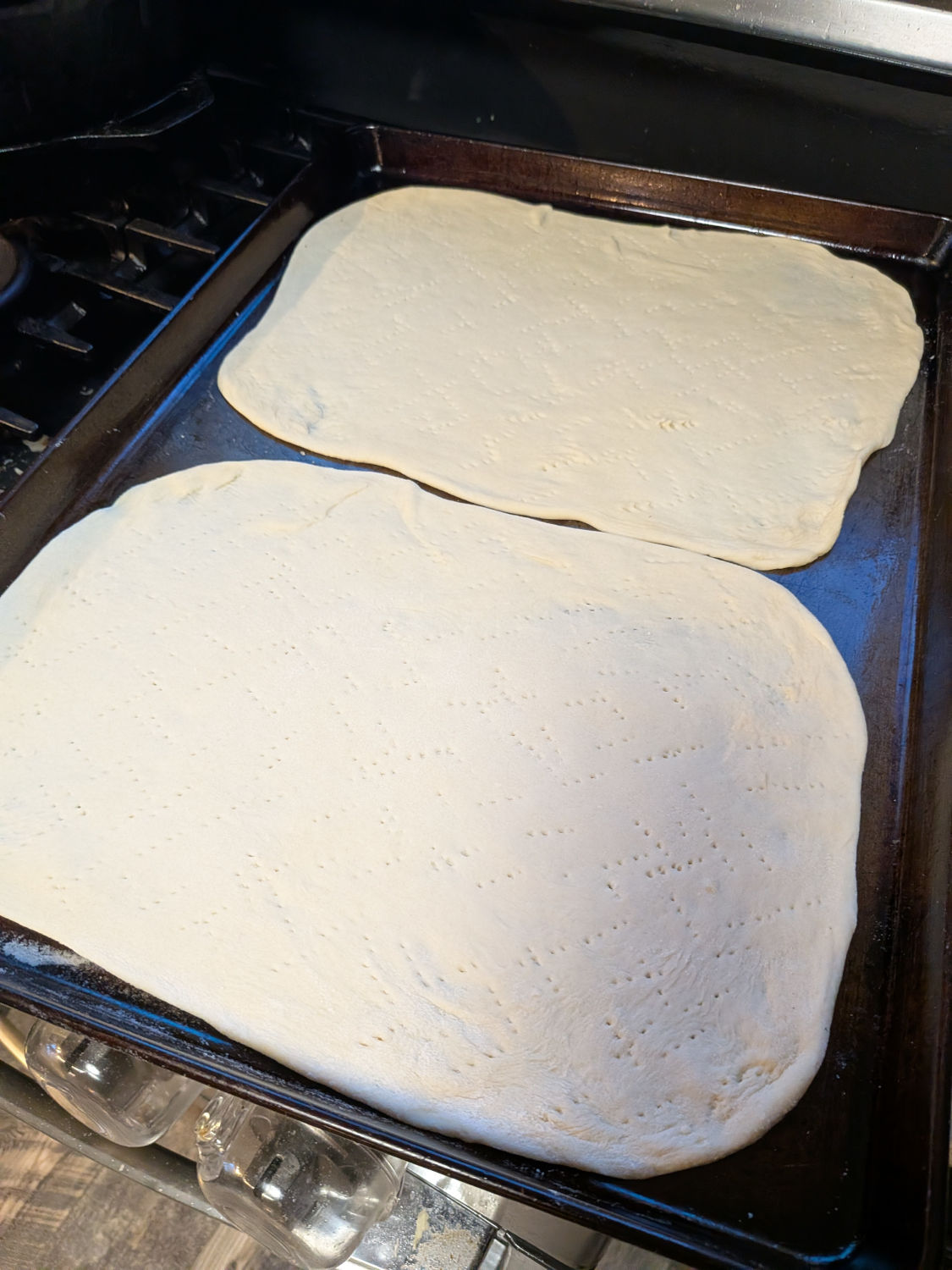 Two tavern-style pizza dough rectangles stretched thin and docked with fork holes on an oiled sheet pan