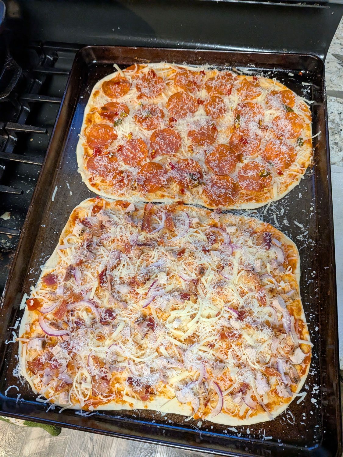 Top-down view of two baked tavern-style sourdough pizzas on a sheet pan, pepperoni-cowboy-candy on top and smoked chicken-bacon-red-onion below