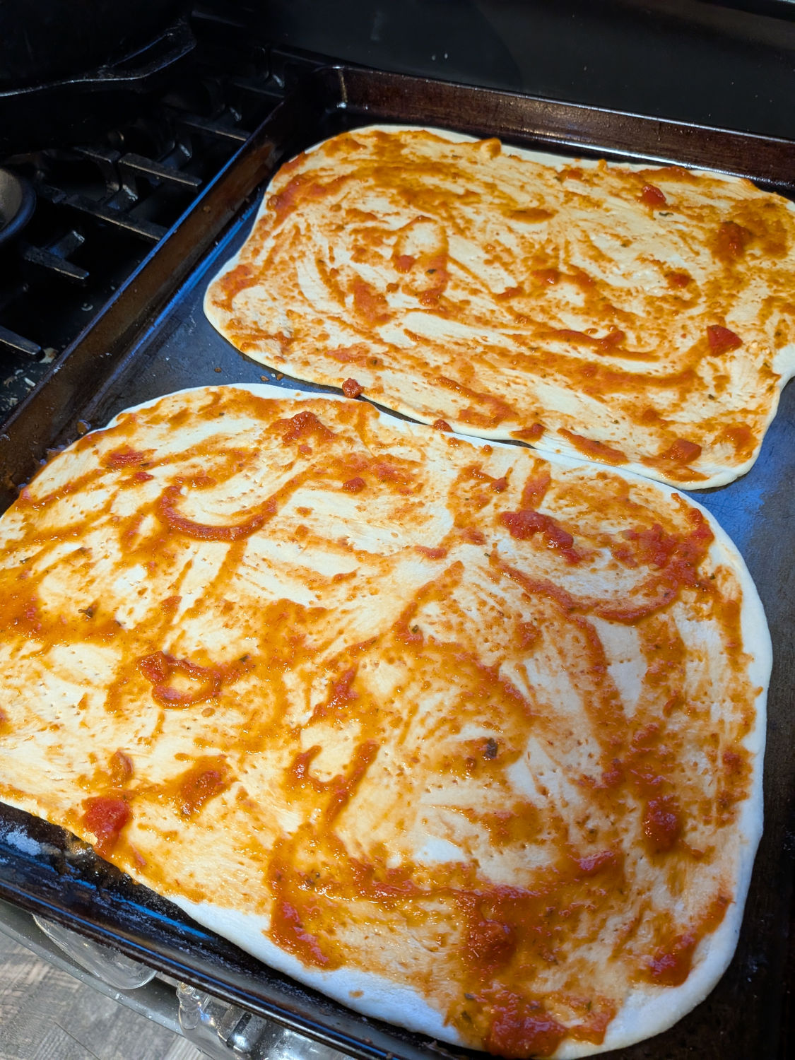 Two stretched tavern-style pizza doughs spread with thin tomato sauce on a sheet pan, before adding cheese and toppings