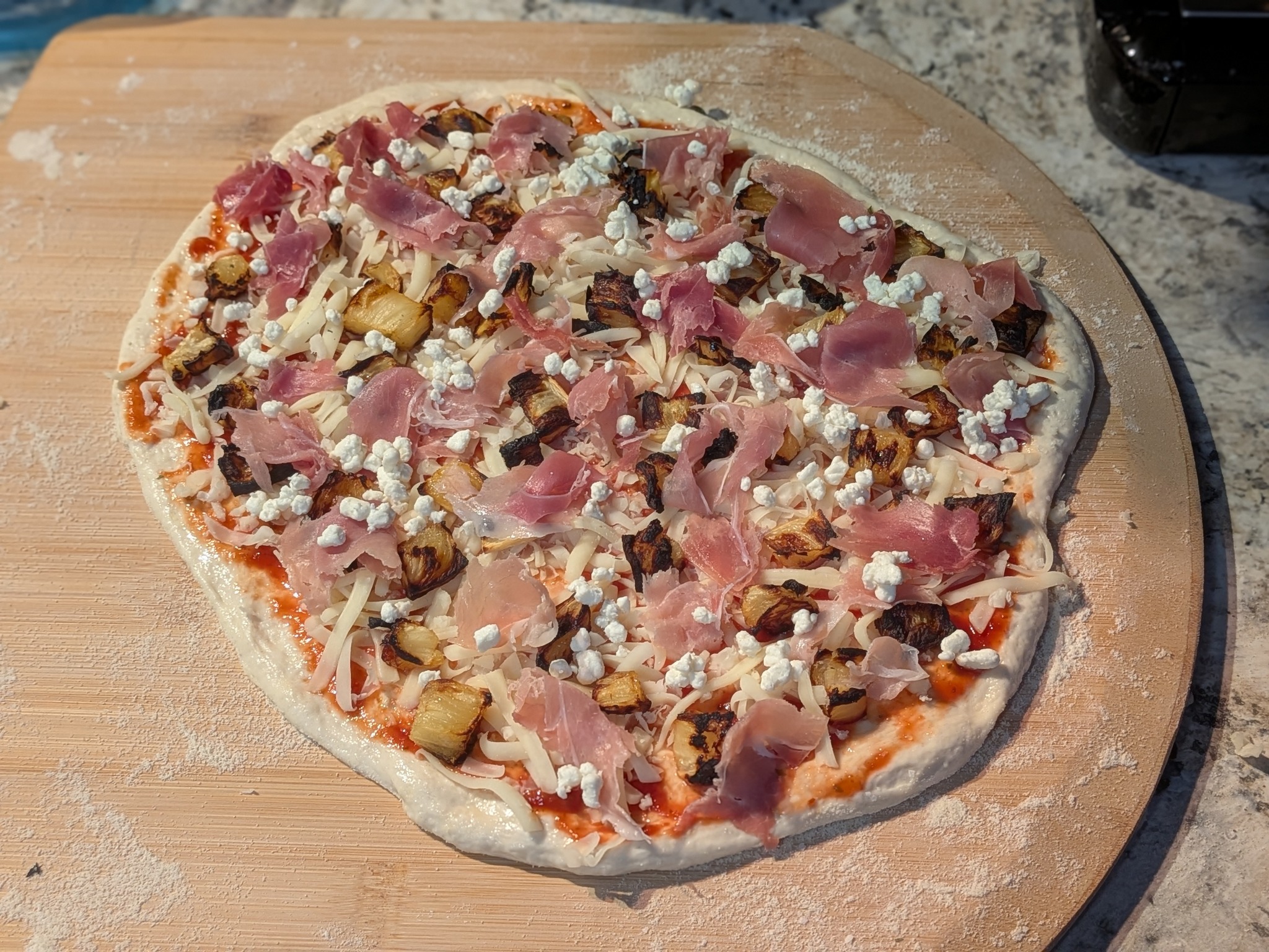 Stretched sourdough Neapolitan pizza topped with prosciutto, already-charred roasted pineapple chunks, and crumbled goat cheese on a peel before baking