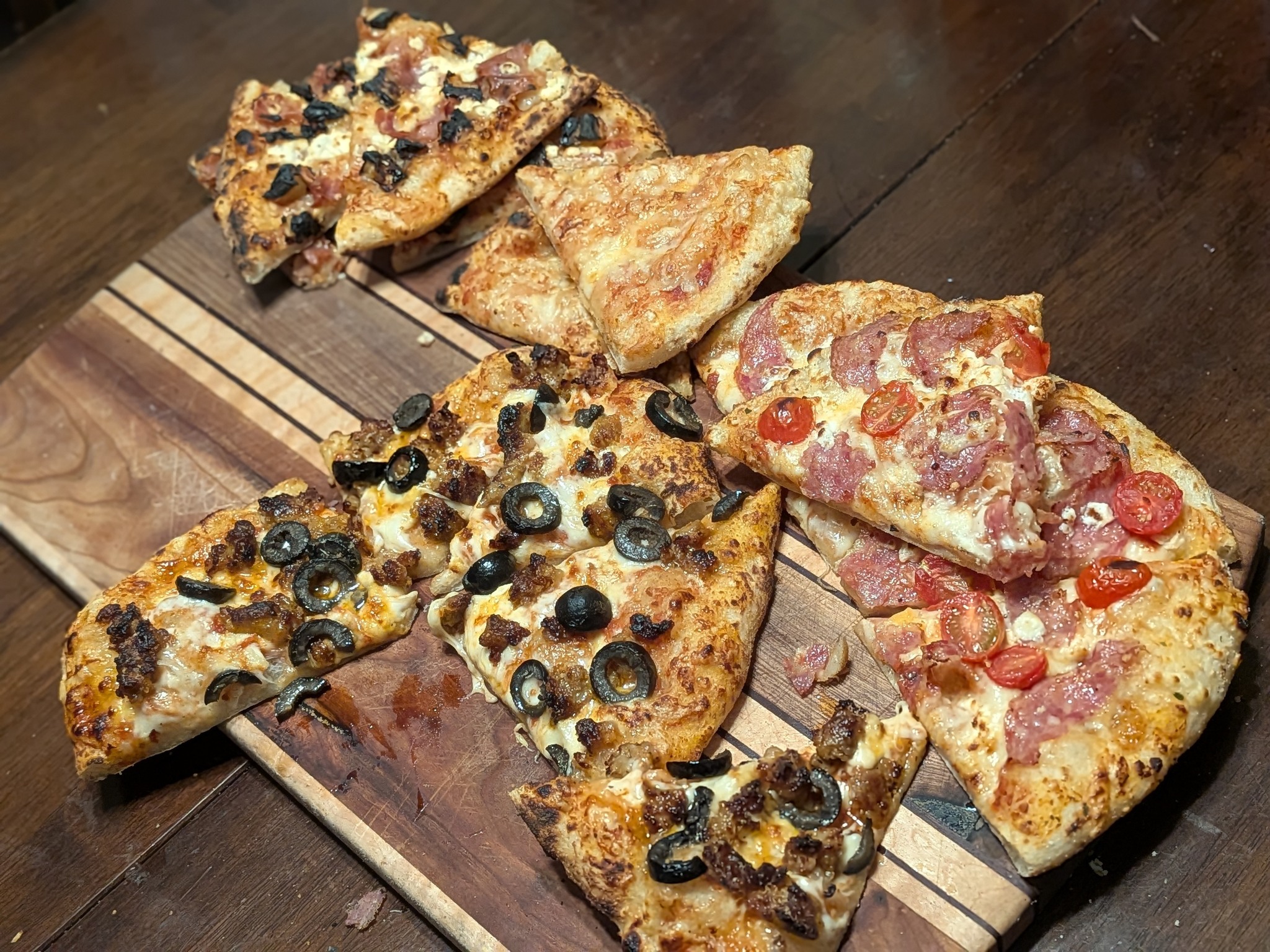 Four cut sourdough Neapolitan pizzas arranged on a wooden cutting board: Italian sausage with black olive, plain cheese, Fancy Hawaiian with prosciutto and pineapple, and salami with cherry tomato and feta