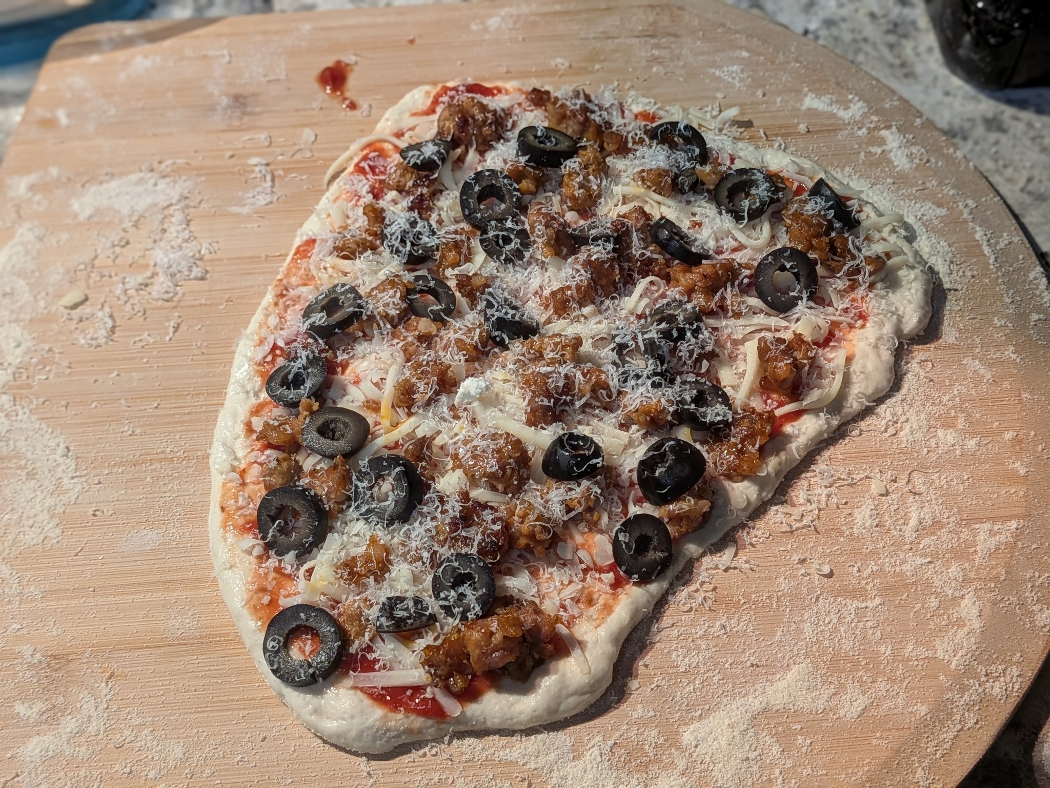 Slightly misshapen sourdough Neapolitan pizza topped with crumbled Italian sausage and sliced black olives on a wooden pizza peel, before baking