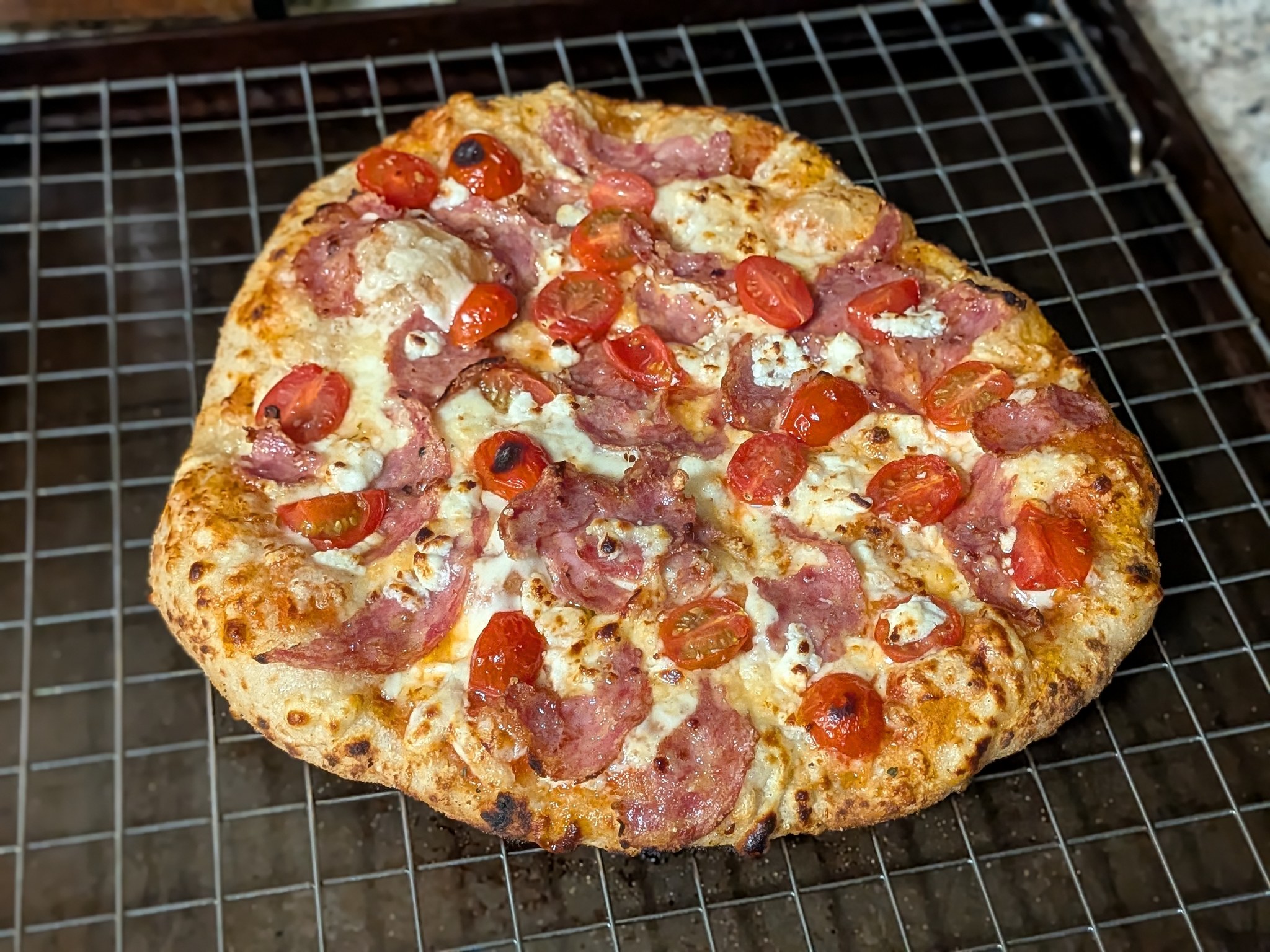 Baked sourdough Neapolitan pizza with salami, halved cherry tomatoes, and crumbled feta on a cooling rack, lightly leoparded crust from the Ooni Koda 16