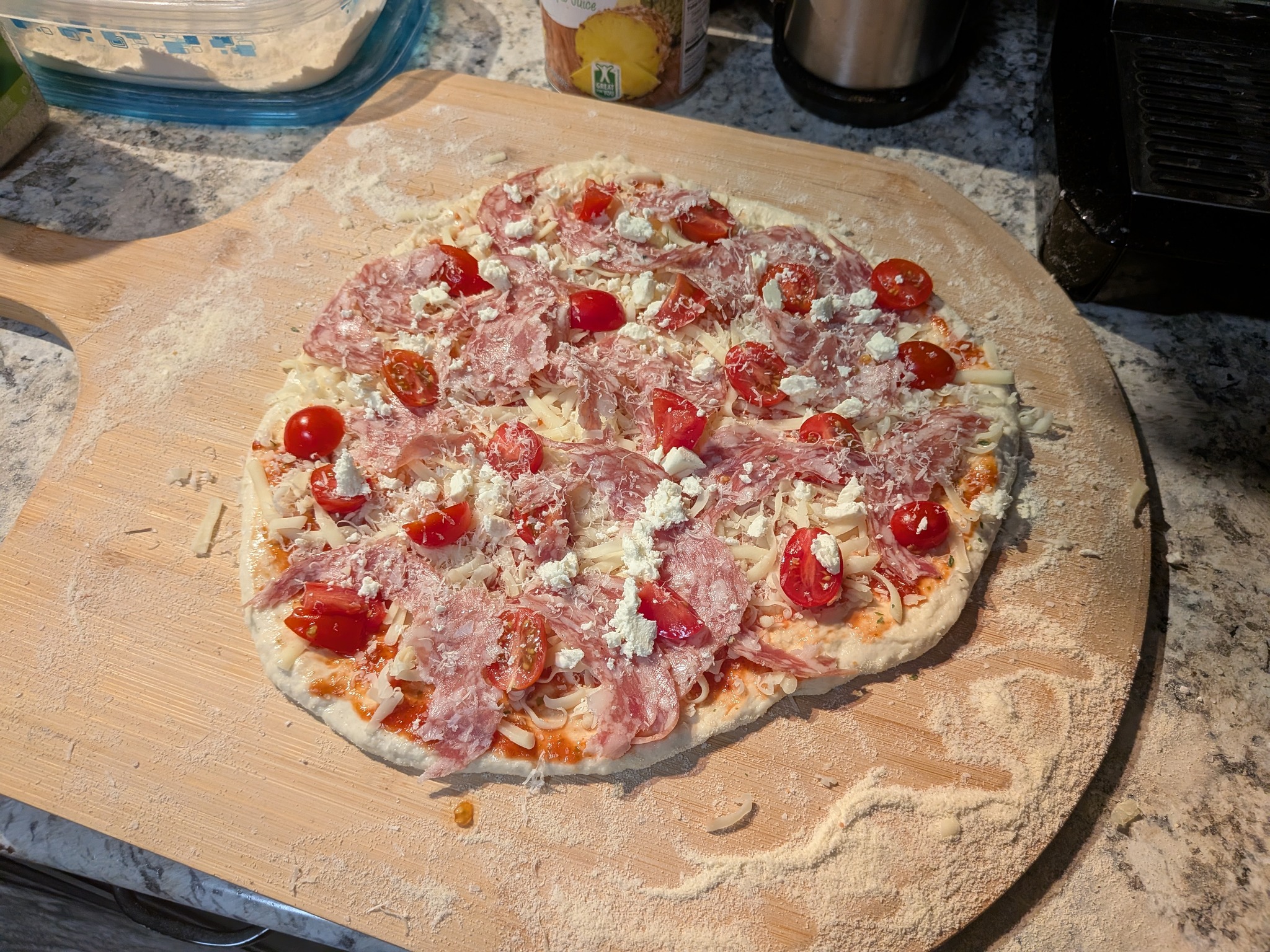 Stretched sourdough Neapolitan pizza topped with salami, halved cherry tomatoes, and crumbled feta on a wooden pizza peel, before going into the Ooni Koda 16