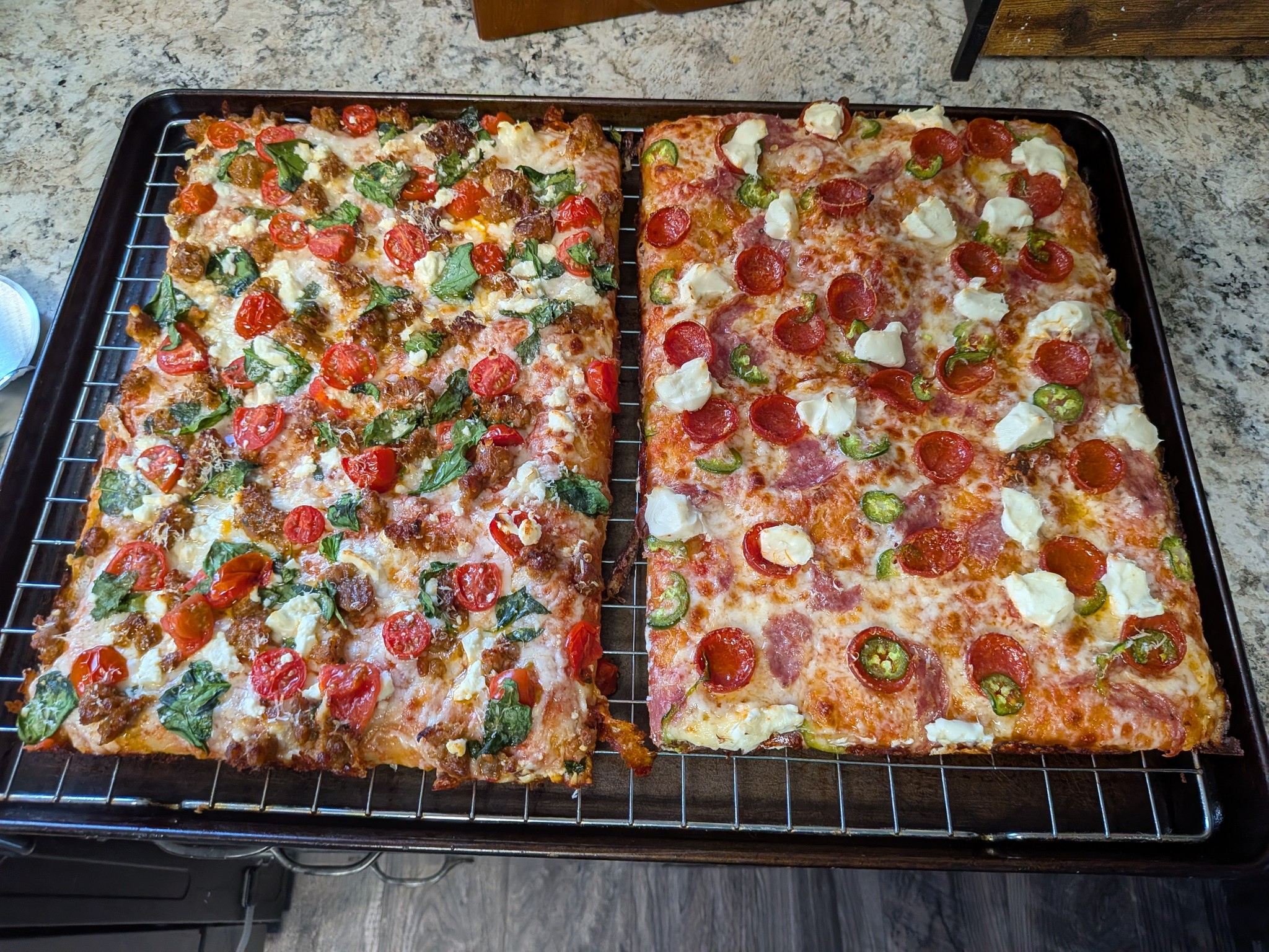 Two cold-fermented Sicilian grandma pan pizzas side by side on a cooling rack, sausage-spinach-cherry tomato-feta on the left and salami-pepperoni-jalapeno-cream cheese on the right