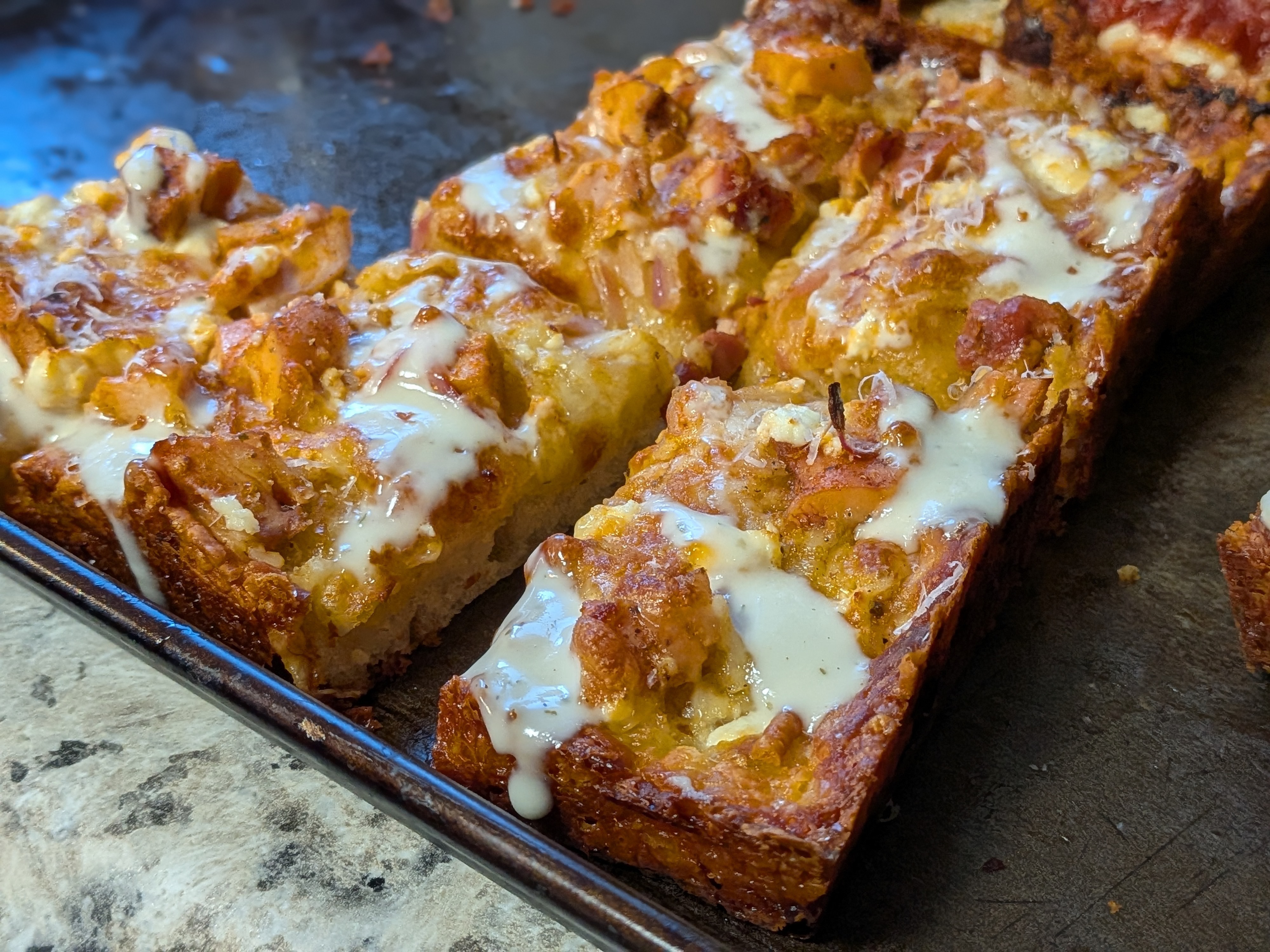 Detroit-style pizza topped with buffalo chicken, red onion, crumbled feta, and a ranch drizzle, deeply caramelized frico cheese edges around the perimeter