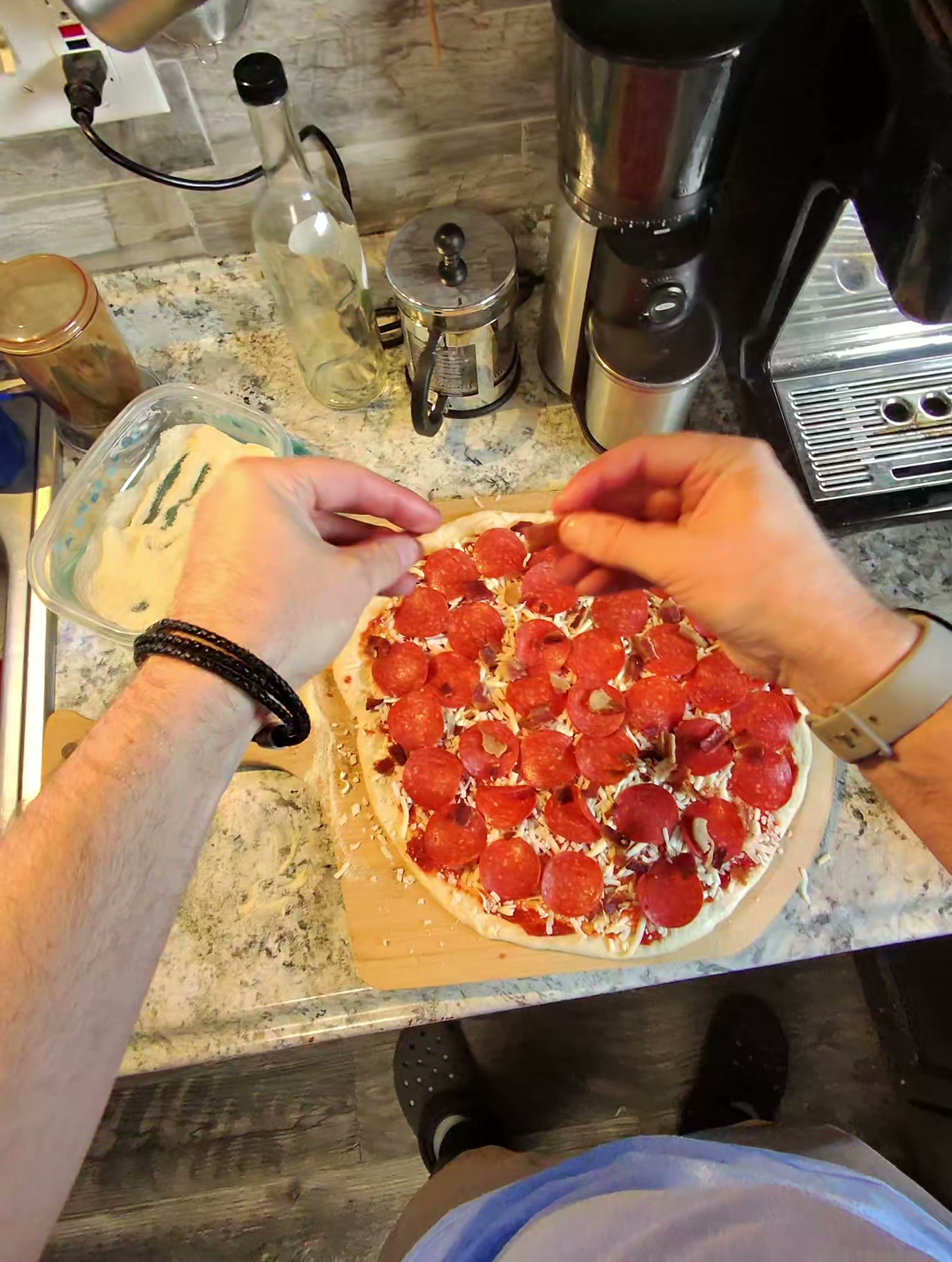 Cold-fermented New York-style pizza topped with pepperoni and bacon on a wooden peel, hands placing the final pepperoni rounds before going into the Ooni Koda 16