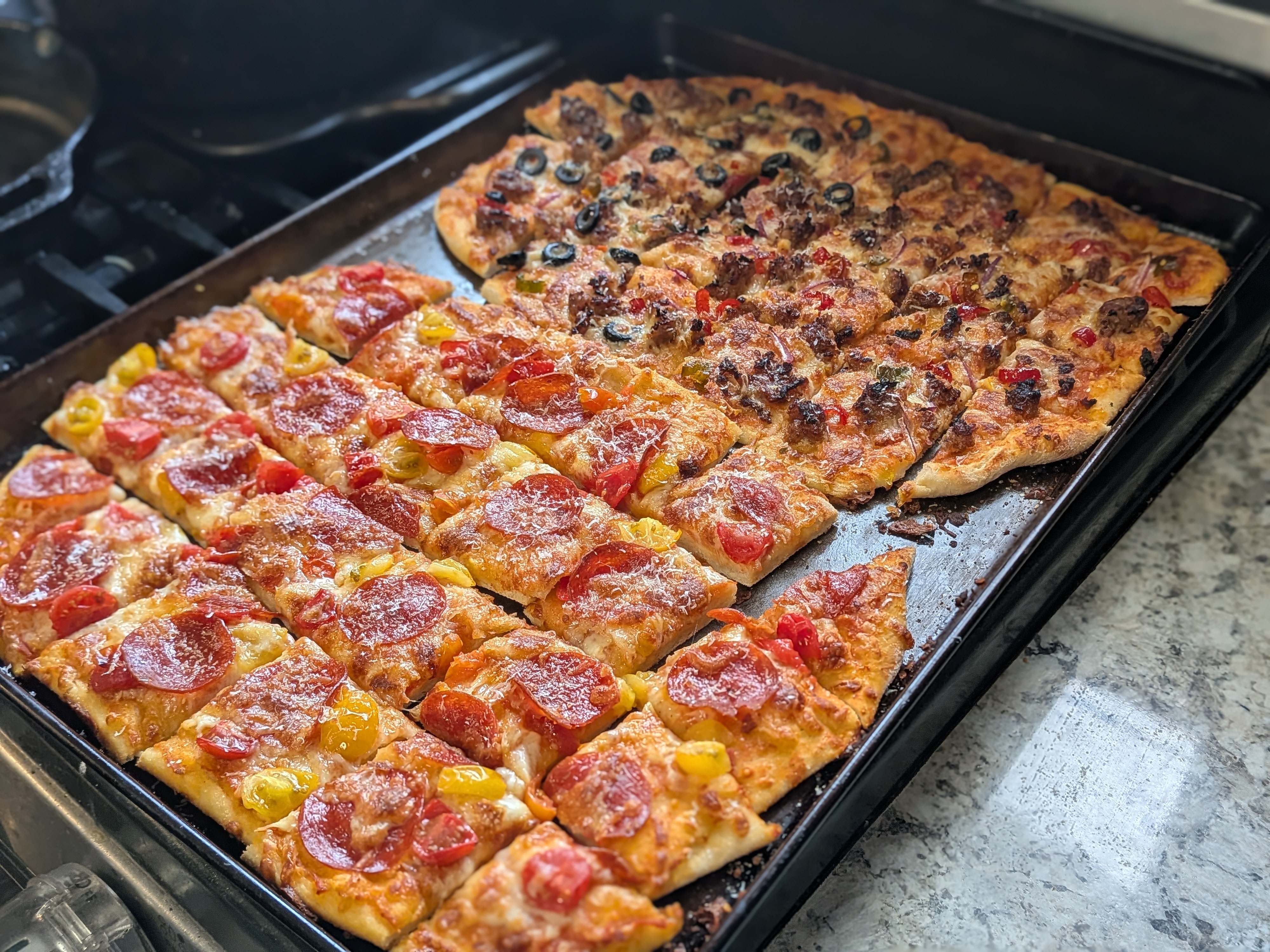 Two large tavern-style thin-crust pizzas on a sheet pan cut into the classic small rectangular squares, pepperoni and cherry tomato in front and Italian sausage with red onion, cowboy candy, and black olives in back