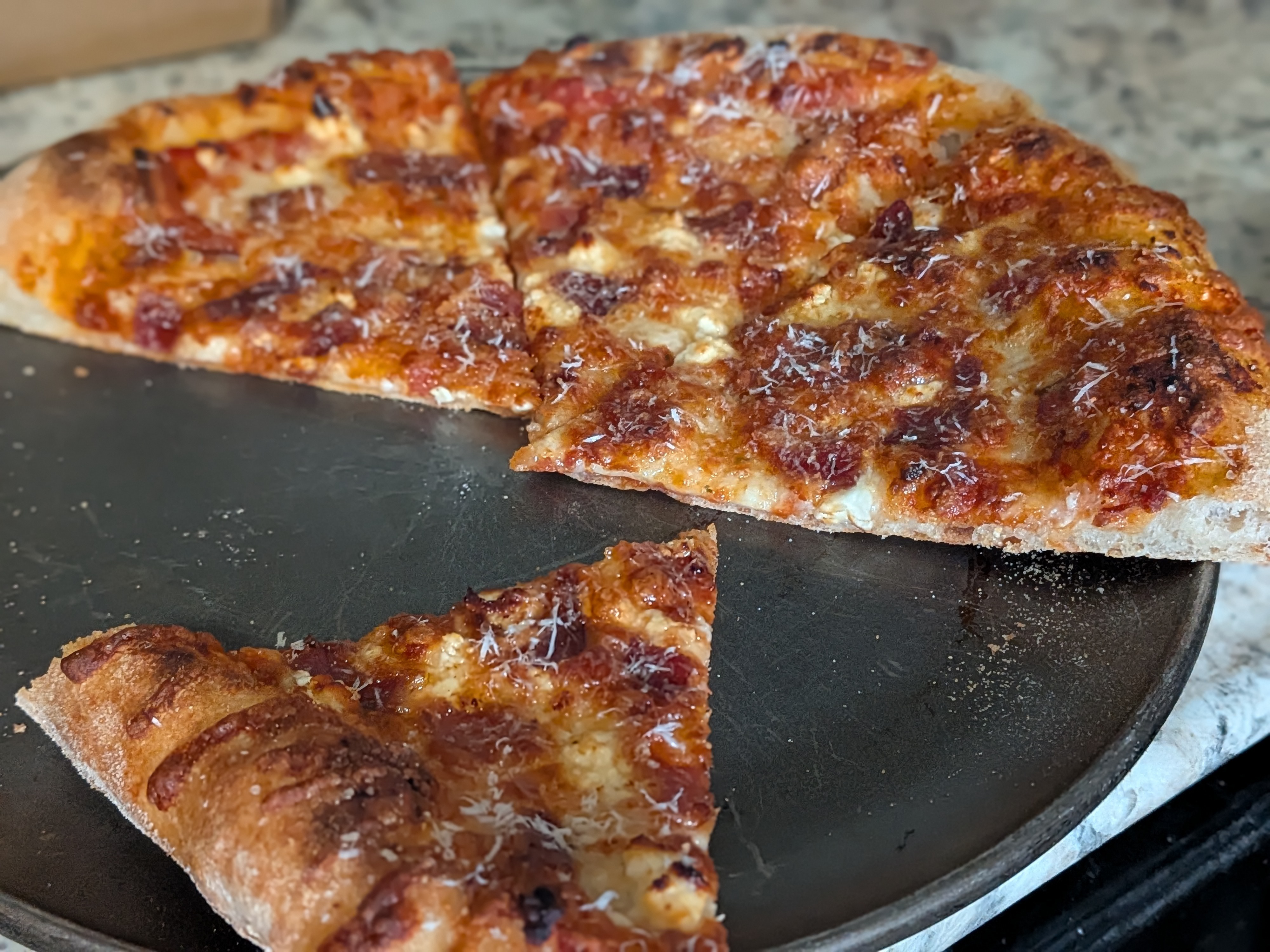 Bacon and feta cheese pizza on a dark pizza pan sliced into wedges with one slice pulled in front, golden bubbled crust and parmesan dusting on top