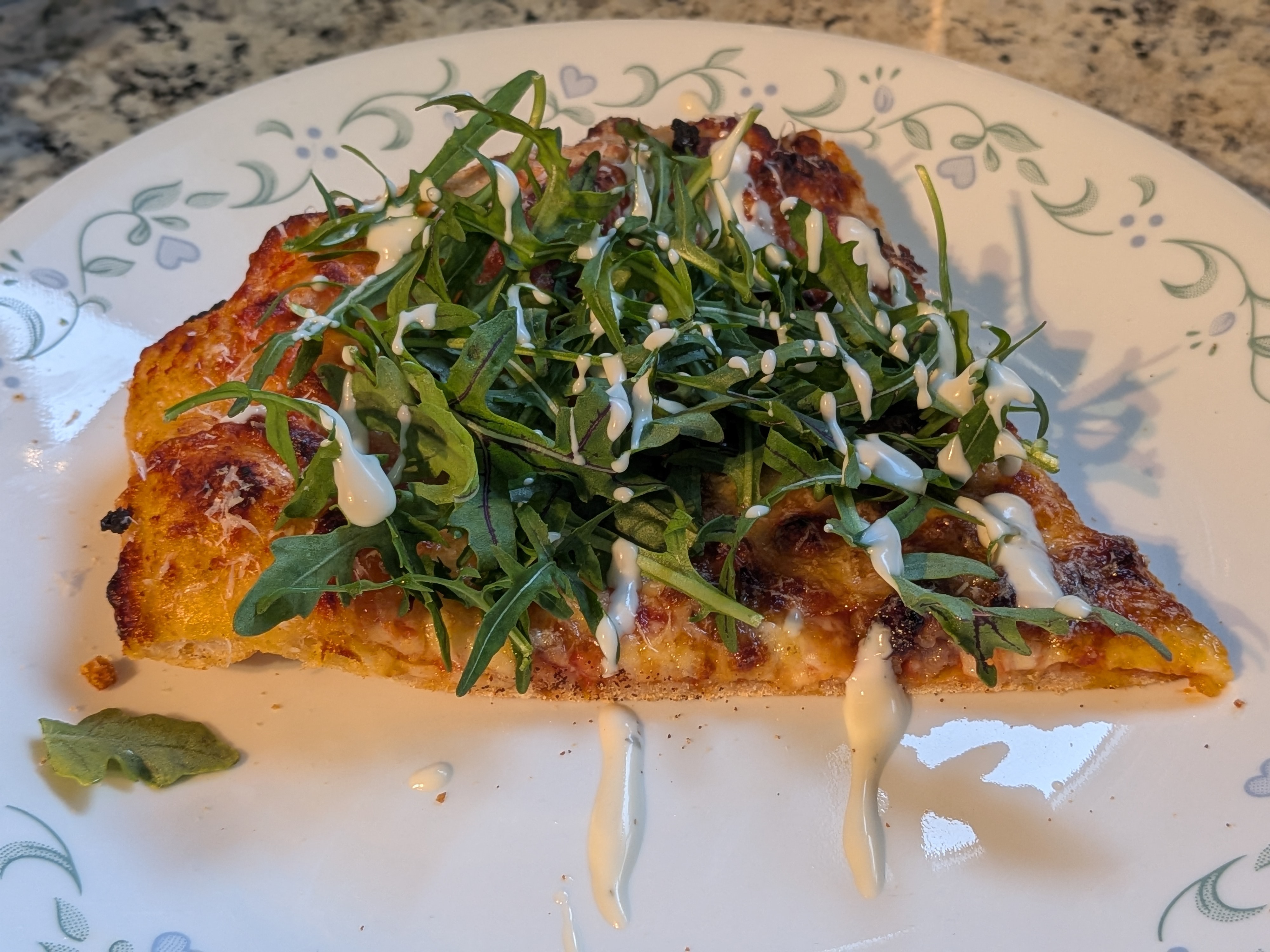 Italian sausage pizza slice topped with fresh arugula and a ranch drizzle on a patterned dinner plate