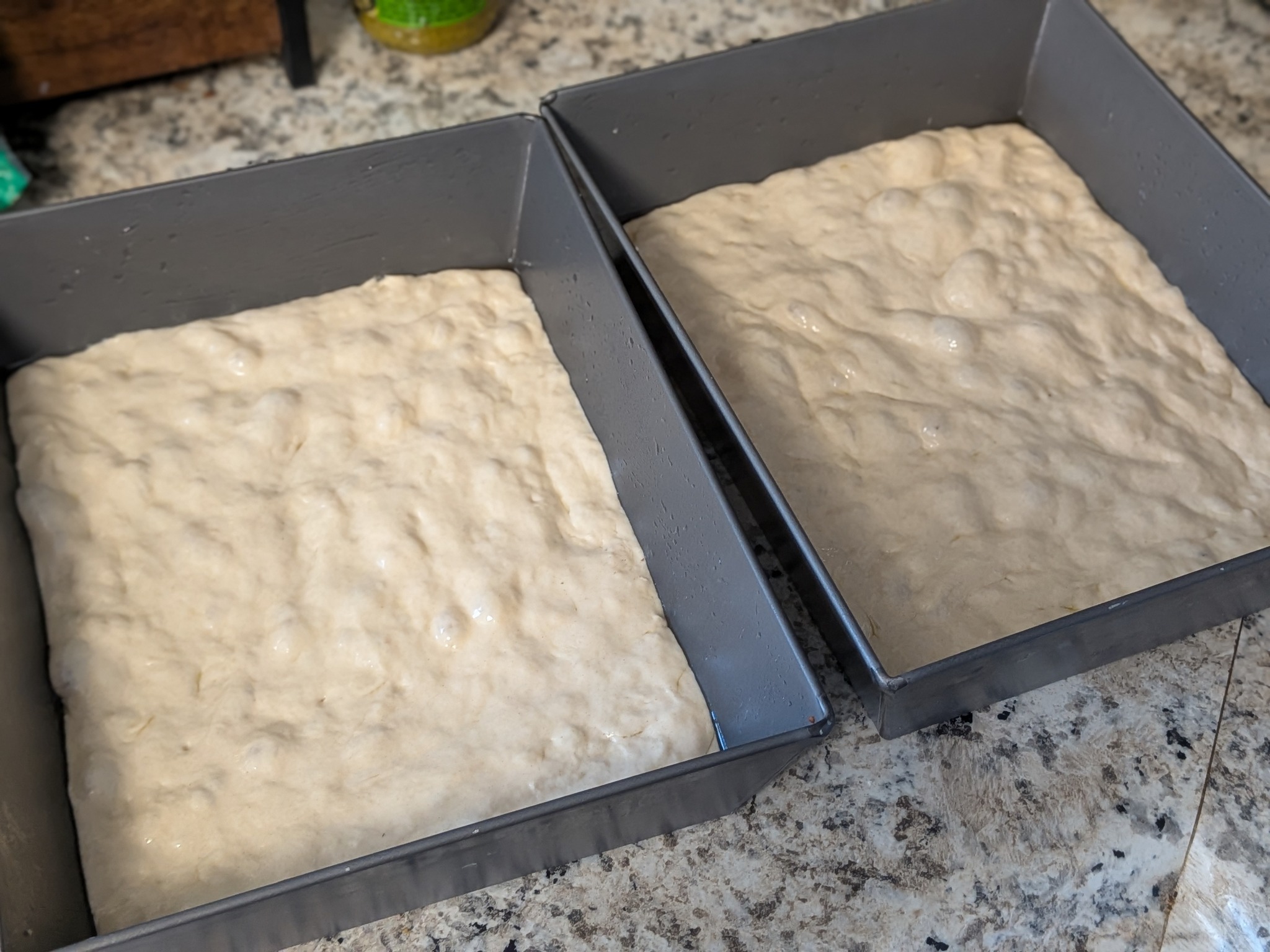 Two pan-proofed dough squares in Detroit-style pans, smooth and dimpled, resting on a granite counter before baking
