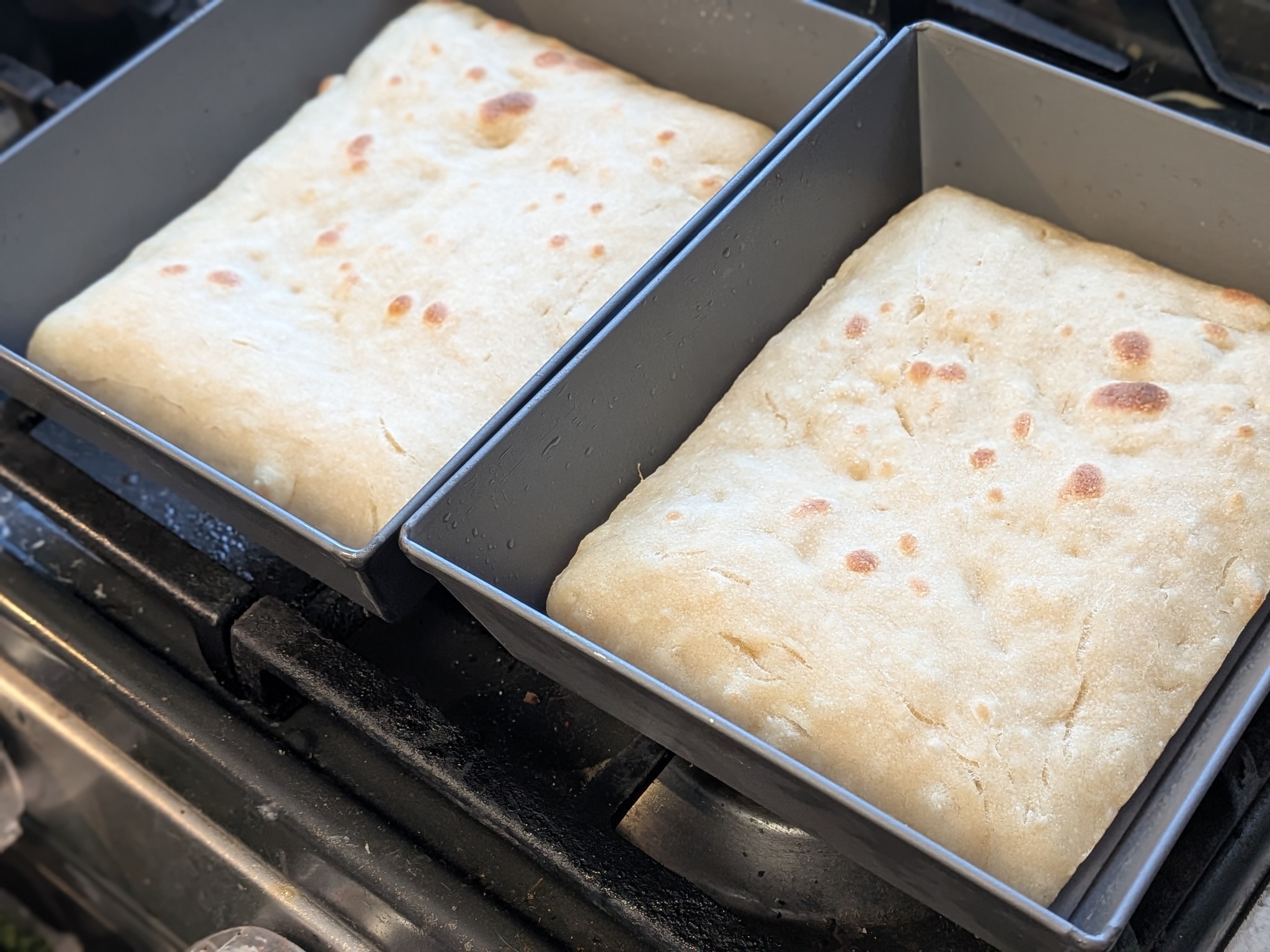 Two par-baked Detroit-style dough squares in pans on the stove, lightly golden with browned blister spots on top