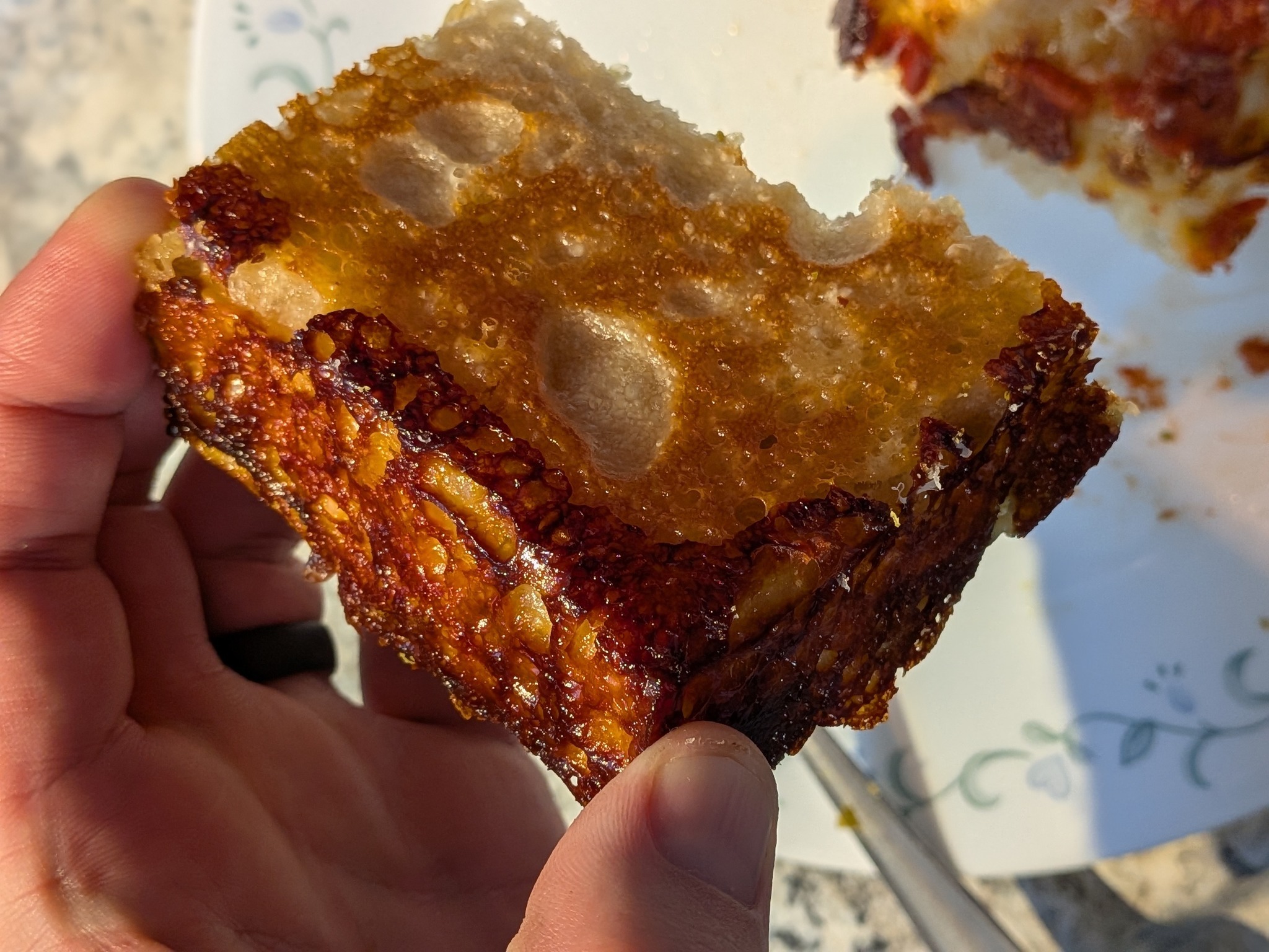 Pepperoni and bacon Detroit pizza slice held in hand, showing the deeply caramelized cheese crust along the bottom and the open crumb above