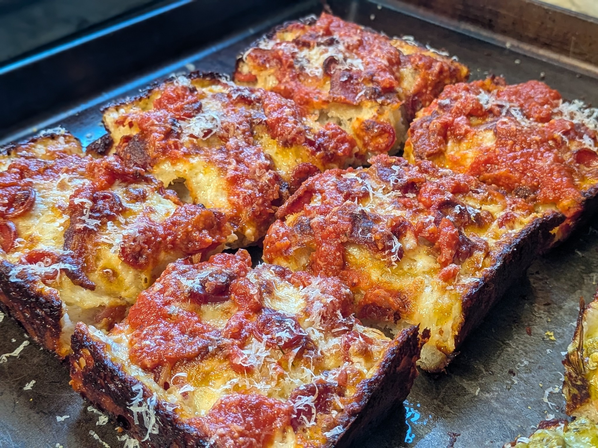 Detroit-style pepperoni and bacon pizza in a dark pan, sliced into squares, with red tomato sauce striped on top and parmesan dusting