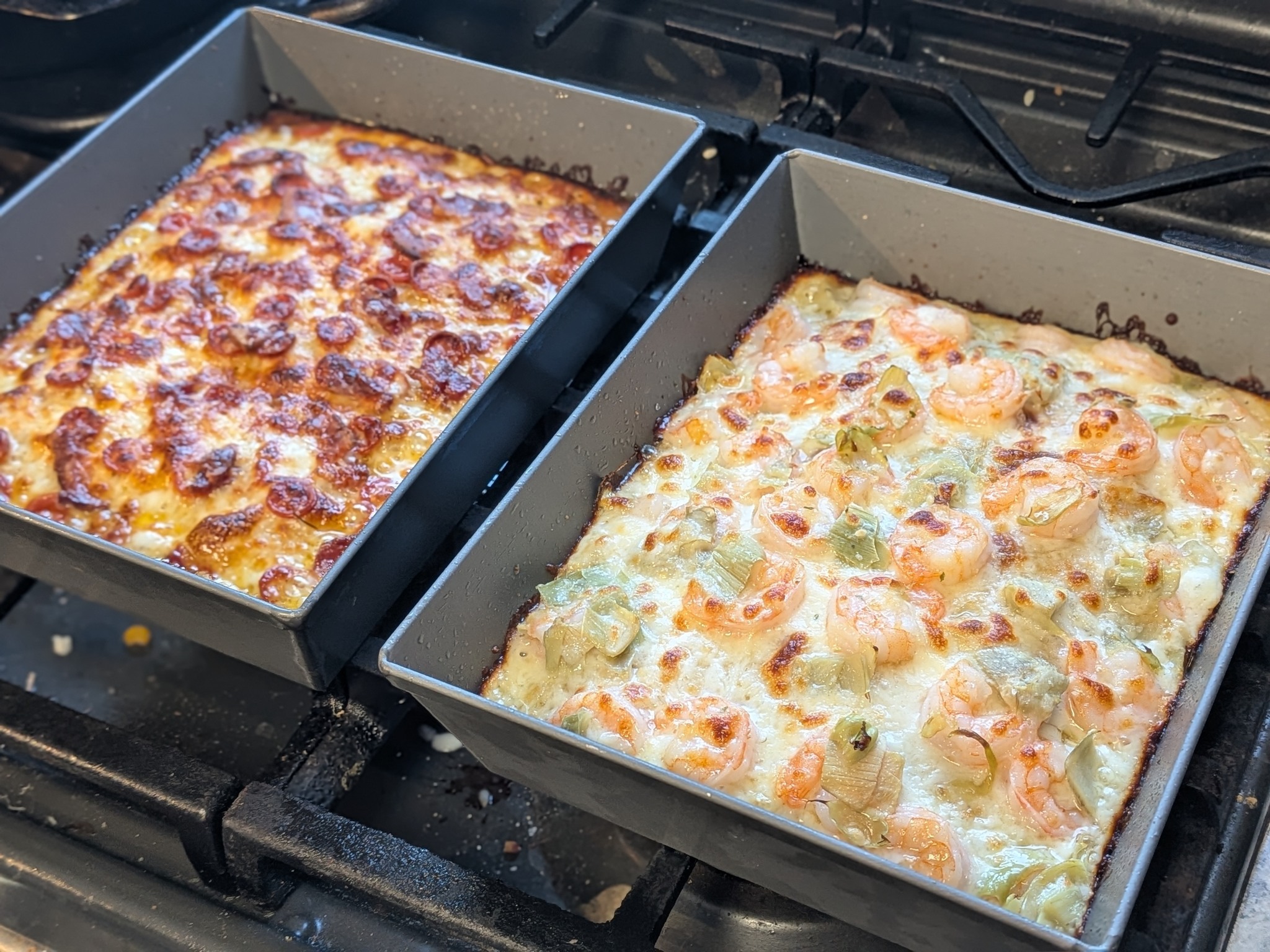 Two Detroit-style pizzas in their pans on the stovetop, pepperoni and bacon at top and shrimp with pesto at bottom, both fresh from the oven