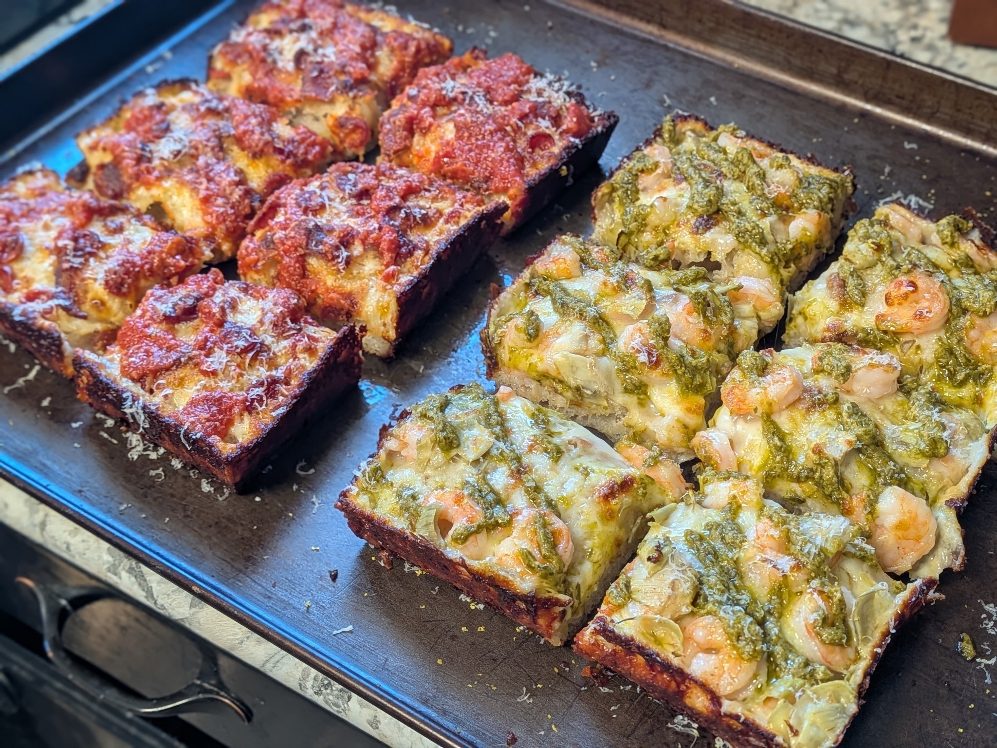 Two Detroit-style pizzas on a dark pan sliced into squares, pepperoni and bacon with tomato sauce stripes on the left, shrimp with basil pesto and artichoke hearts on the right