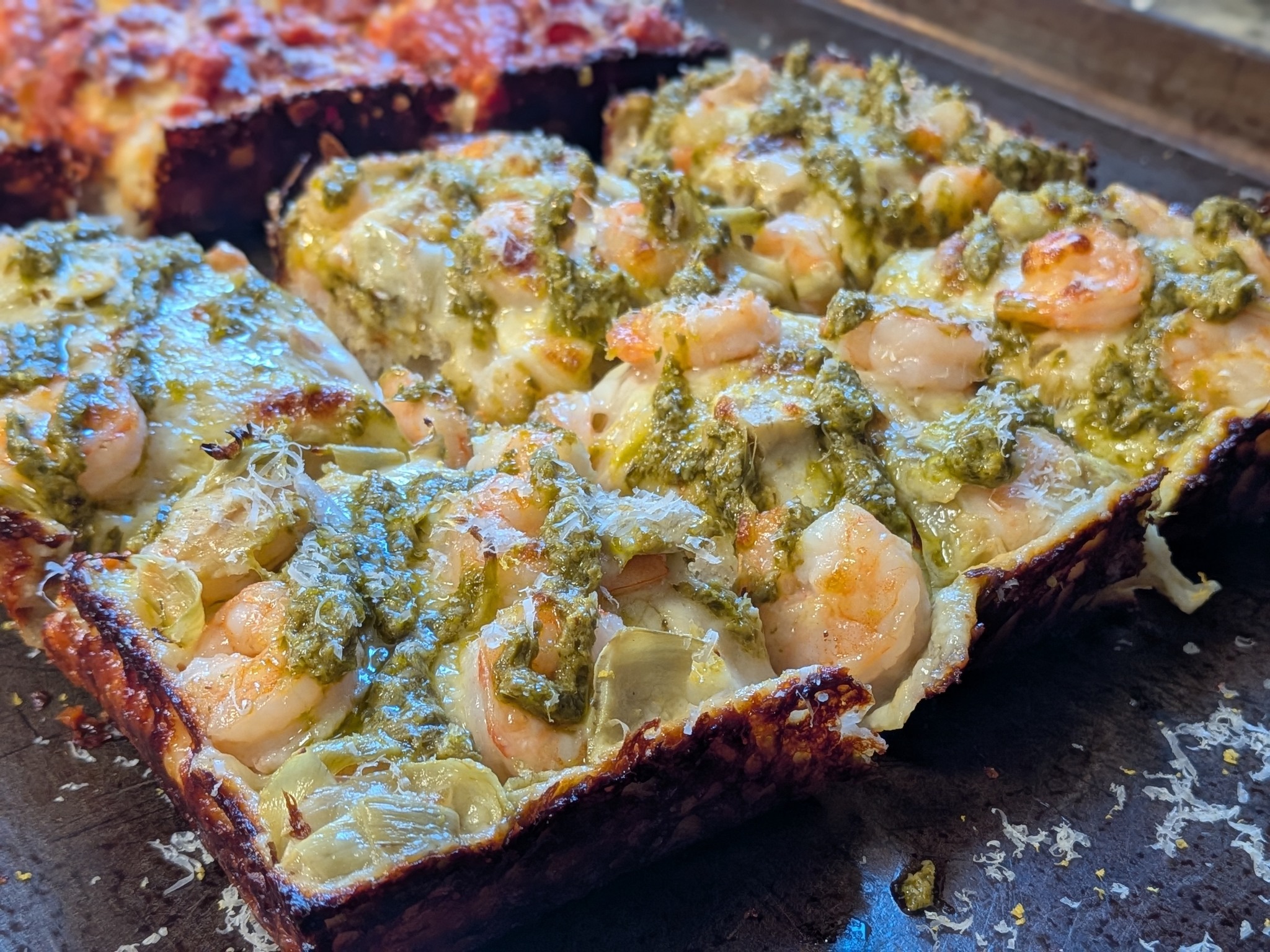 Shrimp, basil pesto, and artichoke heart Detroit pizza in a dark pan, sliced into squares, with pesto dollops and shrimp visible on top